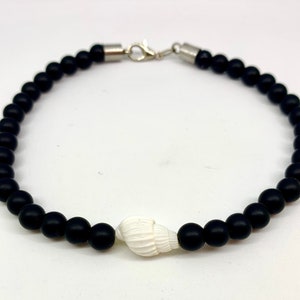 Könnte beinhalten: Ein schwarzes Perlenarmband mit einem weißen Muschel-Anhänger. Das Armband hat einen silbernen Verschluss.
