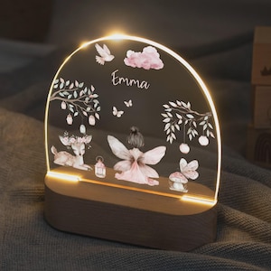 Könnte beinhalten: Dekoratives Nachtlicht mit Holzsockel und klarer Acrylplatte. Die Platte zeigt eine Fee, ein Reh, Schmetterlinge und den Namen "Emma". Das Licht strahlt ein warmes Leuchten aus und erzeugt ein skurriles Ambiente.
