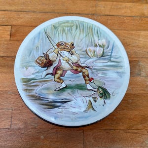 Könnte beinhalten: Eine runde, vintage Blechdose mit einer skurrilen Illustration eines angelnden Frosches. Der Frosch, bekleidet mit einem Mantel, steht auf einem Seerosenblatt und hält eine Angel. Die Szene spielt in einem Teich mit Seerosen und einem Fisch.