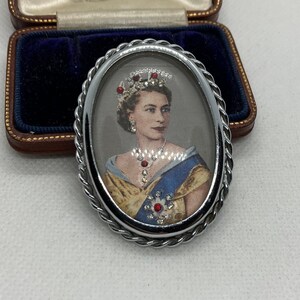 Op de afbeelding: Een ovale broche met een portret van koningin Elizabeth II die een kroon, ketting en blauwe sjerp met rode en doorzichtige stenen draagt. De broche heeft een zilverkleurige, gedraaide metalen rand en zit in een bruin rechthoekig doosje met blauw fluweel.