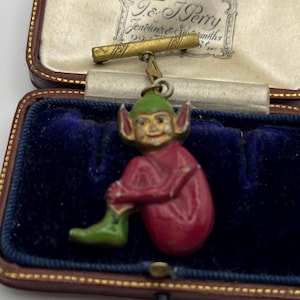 Può includere: Spilla vintage a forma di elfo con corpo rosso, cappello e stivali verdi e viso dorato. La spilla è sospesa a una spilla a barra dorata ed è presentata in una scatola foderata in velluto blu con esterno in pelle marrone.