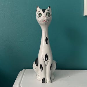 May include: A decorative ceramic cat figurine. The cat is white with black markings, a long neck, and green eyes. It is positioned on a white surface against a teal wall. The figurine is a decorative item.
