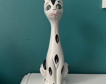 May include: A tall, white ceramic cat figurine with black spots and accents. The cat has a long neck, green eyes, and a red mouth. The figurine is sitting on a white surface against a teal wall.