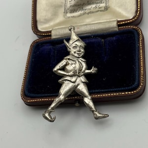 May include: A silver-toned metal brooch shaped like a walking elf with a pointed hat, tunic, and curled-toe shoes. The elf has a smiling expression and is positioned in front of an open, rectangular, dark brown jewelry box with blue velvet lining.