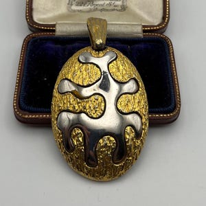 Puede incluir: Colgante dorado con base ovalada texturizada y diseño abstracto plateado. El colgante se exhibe en un joyero vintage con interior de terciopelo azul y exterior de cuero marrón.