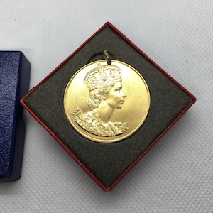 Op de afbeelding: Een goudkleurige medaillon met een profiel van een gekroonde persoon, gepresenteerd in een zwart-rode doos. De medaillon heeft een lus om op te hangen en ligt naast een blauwe doos met gouden tekst.