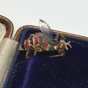 Puede incluir: Broche de abeja dorada con detalles rojos, negros y plateados. La abeja tiene un diseño detallado y un alfiler para sujetar. El broche se encuentra sobre un fondo de terciopelo azul.