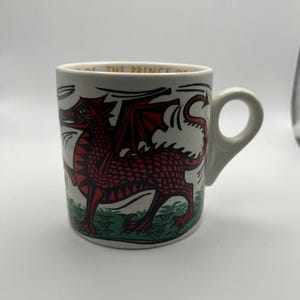 May include: White ceramic mug featuring a red dragon illustration with black accents. The dragon has outstretched wings and is set against a green grassy base. The mug has a curved handle and the text "... OF THE PRINCE ..." is visible.