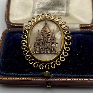 Op de afbeelding: Een antieke goudkleurige broche met een ovale lijst, met een gedetailleerd reliëf van een kathedraal. De broche is geplaatst tegen een crèmekleurige achtergrond en wordt gepresenteerd in een vintage sieradendoos met blauw fluweel.