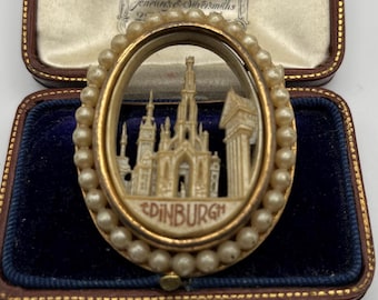 May include: An oval-shaped brooch with a gold-colored frame and a border of small, round pearls. The brooch features a detailed miniature relief of a building with the word "EDINBURGH" at the bottom. The brooch is displayed in a blue velvet-lined box.