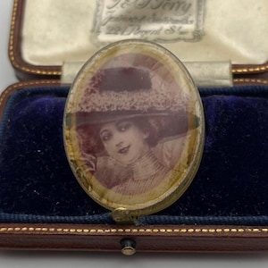 May include: An antique oval-shaped locket with a portrait of a woman in a hat. The locket is set in a gold-toned metal frame and rests in a blue velvet-lined box. The box is brown with a cream-colored satin interior.