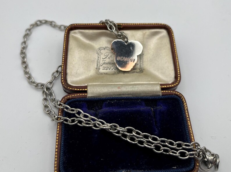 May include: A silver-toned necklace with a Mickey Mouse pendant, displayed in a vintage jewelry box. The pendant features the Disney logo. The box is brown with a blue velvet interior, and the necklace chain is visible.