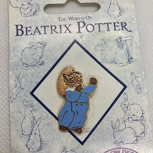 May include: A gold-toned Beatrix Potter pin featuring Peter Rabbit wearing a blue jacket and brown hat. The pin is on a white card with the text "The World of Beatrix Potter" and "High Quality Hand Enamelled Brooch" and the logo "Rainbow Designs".