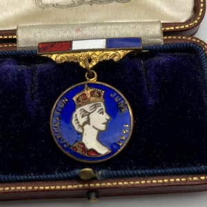 Op de afbeelding: Een vintage kroningsmedaille uit juni 1953, met een profiel van een gekroonde figuur op een blauwe emaille achtergrond. De medaille is opgehangen aan een balk met rood, wit en blauw gestreepte strepen, gepresenteerd in een fluwelen gevoerde koffer.