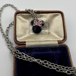 May include: A silver-toned necklace with a Minnie Mouse pendant. The pendant features a black enamel face, black ears, and a pink bow with white polka dots. The necklace is displayed in a vintage jewelry box.
