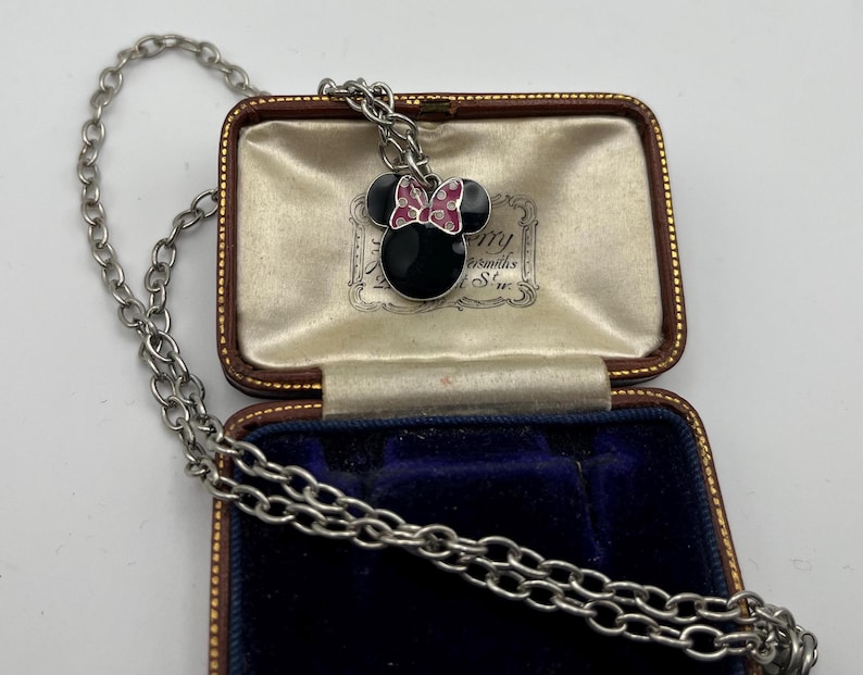 May include: A silver-toned necklace with a Minnie Mouse pendant. The pendant features a black enamel face, black ears, and a pink bow with white polka dots. The necklace is displayed in a vintage jewelry box.