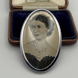 Op de afbeelding: Een ovale broche met een zwart-witportret van koningin Elizabeth II, versierd met kleine doorzichtige strass-steentjes langs de halslijn van de jurk. De broche is gezet in een zilverkleurig metalen frame en gepresenteerd in een bruin doosje.