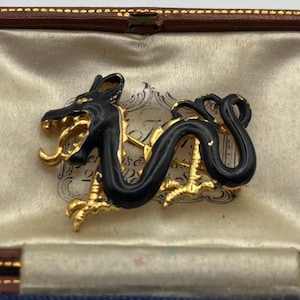 May include: A vintage dragon brooch, black enamel over gold-tone metal. The dragon is in a coiled pose, with detailed scales and a fierce expression. The brooch is presented in a blue and brown jewellery box.