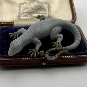 May include: A grey lizard ornament is displayed in a vintage jewellery box with a blue velvet interior. The lizard has detailed scales and is posed as if crawling. The box is brown with gold trim and a cream satin lining.