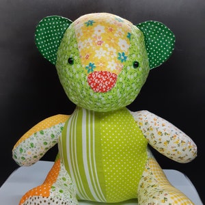 May include: A patchwork green teddy bear made from yellow, orange and white floral fabric. The bear has green ears with white polka dots and black button eyes.