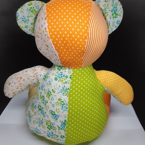 May include: A patchwork teddy bear made from orange, green and white fabric. The bear has a floral pattern on its ears and is sitting on a white surface.