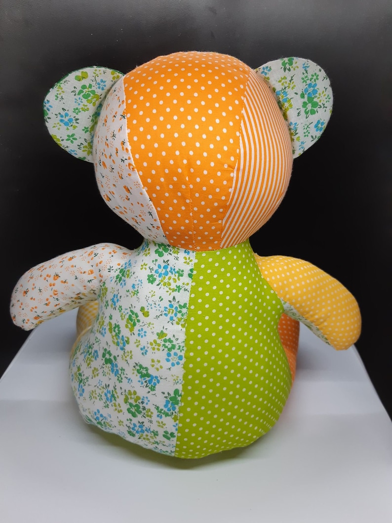 May include: A patchwork teddy bear made from orange, green and white fabric. The bear has a floral pattern on its ears and is sitting on a white surface.