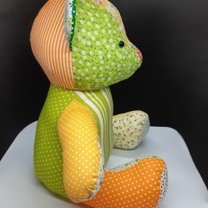 May include: A patchwork teddy bear made of green, orange, yellow and white fabric. The bear has a green and white striped body, a green and white floral head, and orange and white polka dot legs.