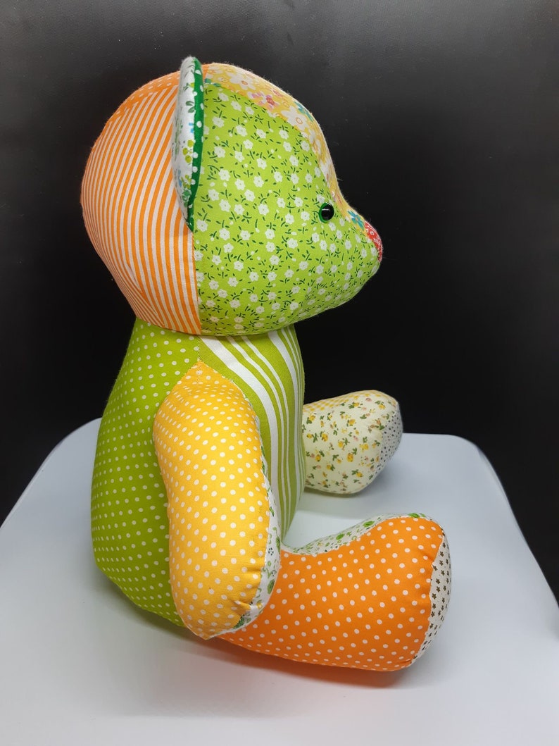 May include: A patchwork teddy bear made of green, orange, yellow and white fabric. The bear has a green and white striped body, a green and white floral head, and orange and white polka dot legs.