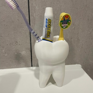 Puede incluir: Un soporte blanco para cepillos de dientes con forma de diente con un cepillo de dientes morado, un cepillo de dientes amarillo con un diseño de dibujos animados y un tubo de pasta de dientes "SYSTEMA". El soporte está sobre una superficie blanca.