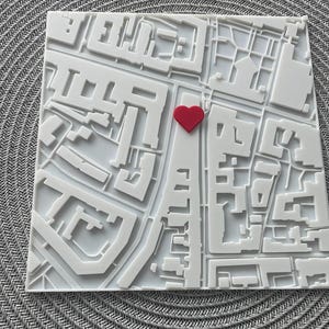 May include: A white, three-dimensional map with a red heart accent. The map depicts a city layout with buildings and streets. The heart is centrally placed, adding a touch of color to the otherwise monochromatic design. The map is square-shaped.