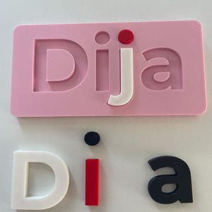 Op de afbeelding: Een roze rechthoekig bord met het woord "Dija" in witte, rode en zwarte letters. De letters zijn los en kunnen worden gerangschikt. De letters "Dija" staan in een roze kader. De letters "Dia" zijn ook los.