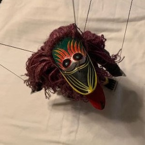 May include: A colorful wooden puppet with a red, yellow, green, and black face mask. The puppet has long, pink yarn hair and is attached to strings.