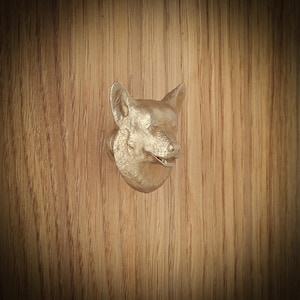 May include: A gold-coloured fox head door knob mounted on a wooden surface. The fox head is detailed with visible ears, nose, and mouth. The wood grain is visible in the background.