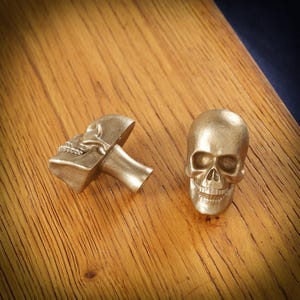 May include: Two gold-colored skull-shaped cabinet knobs. One knob is whole, while the other is cut in half, revealing the mounting stem. The knobs are made of metal and rest on a wooden surface.