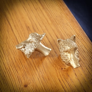 May include: Two gold-coloured cabinet knobs, each shaped like a detailed boar's head. The knobs are set against a wooden surface, showcasing their metallic finish and intricate design. These decorative knobs add a unique touch to furniture.