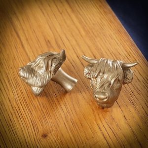 May include: Two metallic gold-colored Highland cow head cufflinks. The cufflinks are detailed with horns, fur, and facial features. They are placed on a wooden surface.