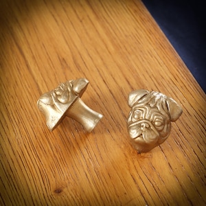 May include: Two gold-coloured cabinet knobs, one shaped like a pug's head and the other with a mushroom-like design. The knobs are on a wooden surface, highlighting their detailed features and metallic sheen.