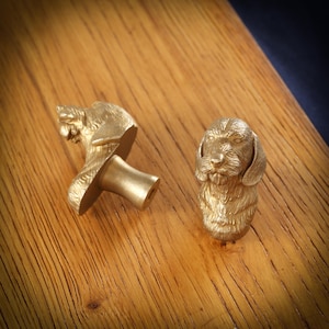 May include: Two gold-coloured dog-shaped cabinet knobs. One knob is detached, showing the stem for attachment. The other knob is upright. The knobs are made of metal and are on a wooden surface.