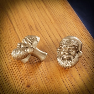 May include: Two bronze-coloured Santa Claus cabinet knobs. One knob is a full Santa face, and the other is a side profile. The knobs are detailed with a hat, beard, and smiling expression. The knobs are on a wooden surface.