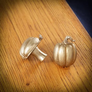 Puede incluir: Dos tiradores dorados para armarios. Uno tiene forma de seta, el otro de calabaza. El tirador de seta tiene un pequeño detalle de hoja. El tirador de calabaza tiene un tallo curvado. Ambos están sobre una superficie de madera.