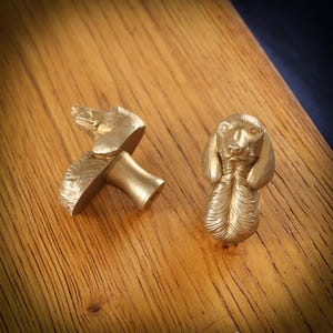 May include: Two brass-colored dog-shaped cabinet knobs. One knob is a full dog figure, the other is a dog head with a stem. The knobs are on a wooden surface.