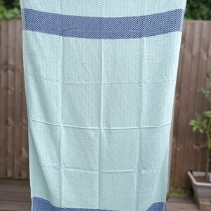 May include: A light blue beach towel with a herringbone pattern and two navy blue stripes. The towel has white fringe on the edges.