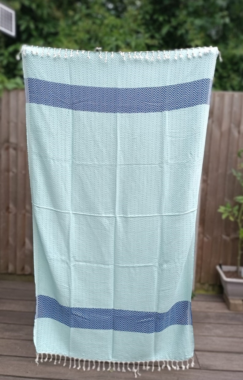 May include: A light blue beach towel with a herringbone pattern and two navy blue stripes. The towel has white fringe on the edges.