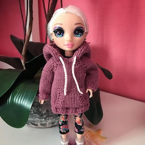 May include: A doll wearing a mauve knitted hoodie with a white drawstring and floral leggings.