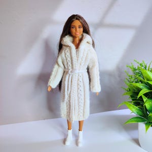 May include: A doll wearing a white, cable-knit robe with a matching belt and white boots. The robe has a shawl collar and is knee-length. The doll has long, dark hair and is standing in front of a light background.