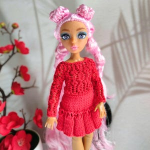 May include: A fashion doll with pink hair styled in two buns and long flowing locks. The doll wears a red knitted sweater and skirt. The doll has blue eyes and freckles. The background is a soft, neutral color with a floral accent.