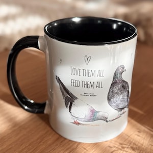 Könnte beinhalten: Ein weißer Keramikbecher mit schwarzem Innenraum und Henkel. Die Außenseite zeigt zwei Taubenillustrationen, drei kleine Herzumrisse und den Text: LOVE THEM ALL FEED THEM ALL. Darunter steht in kleinerer Schrift: Kein Tier verdient Hunger.