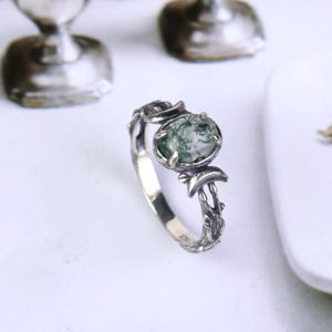 Puede incluir: Un anillo de plata con una piedra ovalada de ágata musgosa. El anillo presenta un diseño inspirado en la naturaleza con detalles de hojas y ramas. La piedra es una mezcla de verde y blanco.