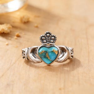 May include: A silver Claddagh ring with a turquoise heart-shaped stone and a crown. The ring features hands clasping the heart. The turquoise stone has gold flecks. The ring is on a wooden surface.