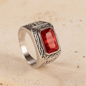 Puede incluir: Un anillo de plata con una gema roja rectangular. El anillo tiene un diseño ornamentado alrededor de la gema y en la banda. La gema es de un color rojo vibrante, y el anillo parece ser de plata de ley.